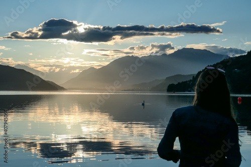 A woman standing at the shore of Millstaetter lake during the sunset. High Alps around. The sun sets behind thick clouds. Calm surface of the lake reflects the orange sky and the mountains. Day end photo