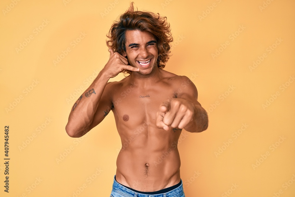 Fototapeta premium Young hispanic man standing shirtless smiling doing talking on the telephone gesture and pointing to you. call me.