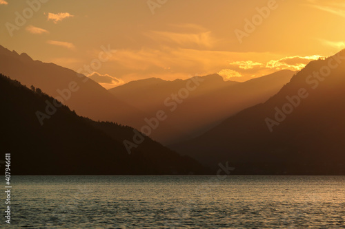 A sunset by Millstaetter lake in Austria. The lake is surrounded by high Alps. Calm surface of the lake reflecting the sunbeams. The sun sets behind the mountains. A bit of overcast. Natural beauty © Chris