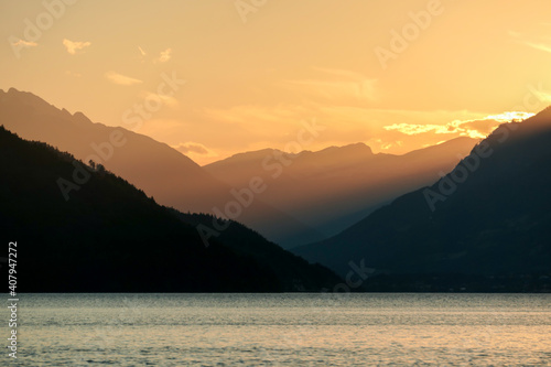 A sunset by Millstaetter lake in Austria. The lake is surrounded by high Alps. Calm surface of the lake reflecting the sunbeams. The sun sets behind the mountains. A bit of overcast. Natural beauty