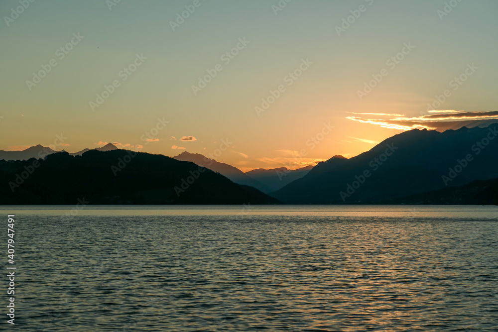 A sunset by Millstaetter lake in Austria. The lake is surrounded by high Alps. Calm surface of the lake reflecting the sunbeams. The sun sets behind the mountains. A bit of overcast. Natural beauty