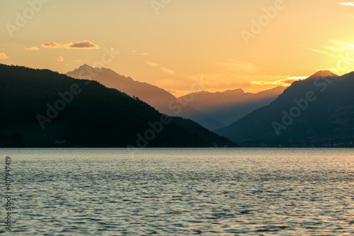 A sunset by Millstaetter lake in Austria. The lake is surrounded by high Alps. Calm surface of the lake reflecting the sunbeams. The sun sets behind the mountains. A bit of overcast. Natural beauty © Chris