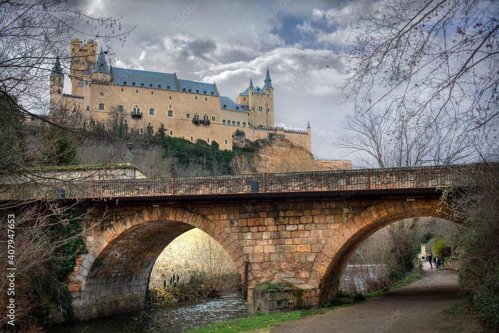 Primer plano de un puente y al fondo tenemos el Alcazar Segovia y a lo lejos dos turistas fotografiando el Alcazar.