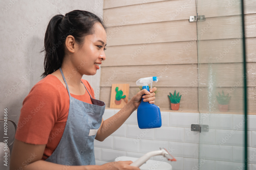 Smiling women spray using a bottle sprayer and hold the window wipers while cleaning the glass in the bathroom Smiling women spray using a bottle sprayer and hold the window wipers while cleaning the glass in the bathroom