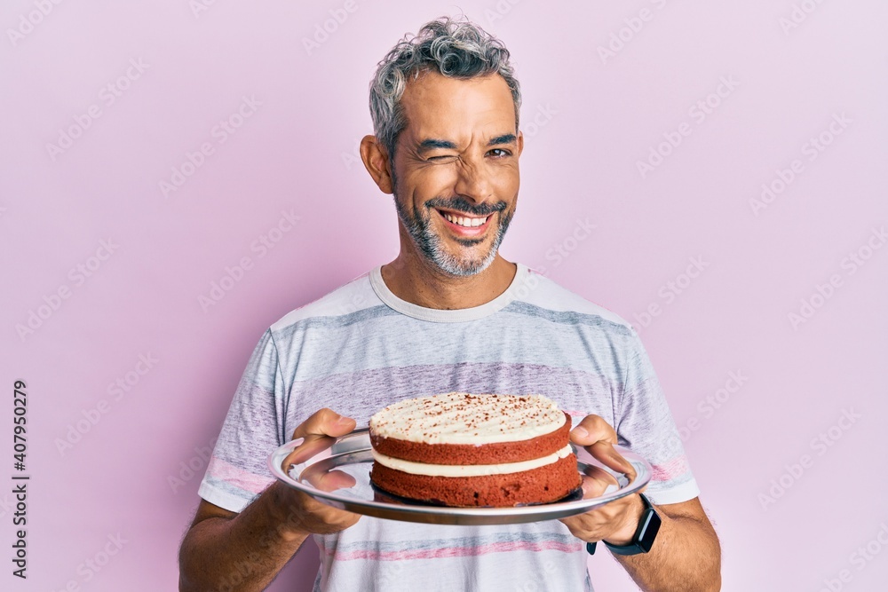 Middle age grey-haired man holding carrot cake winking looking at the camera with sexy expression, cheerful and happy face. Middle age grey-haired man holding carrot cake winking looking at the camera with sexy expression, cheerful and happy face.