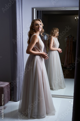Young beautiful blonde girl wearing a full-length silver white chiffon prom ball gown decorated with sparkles and sequins. Model in front of mirror in a fitting room at dress hire service.