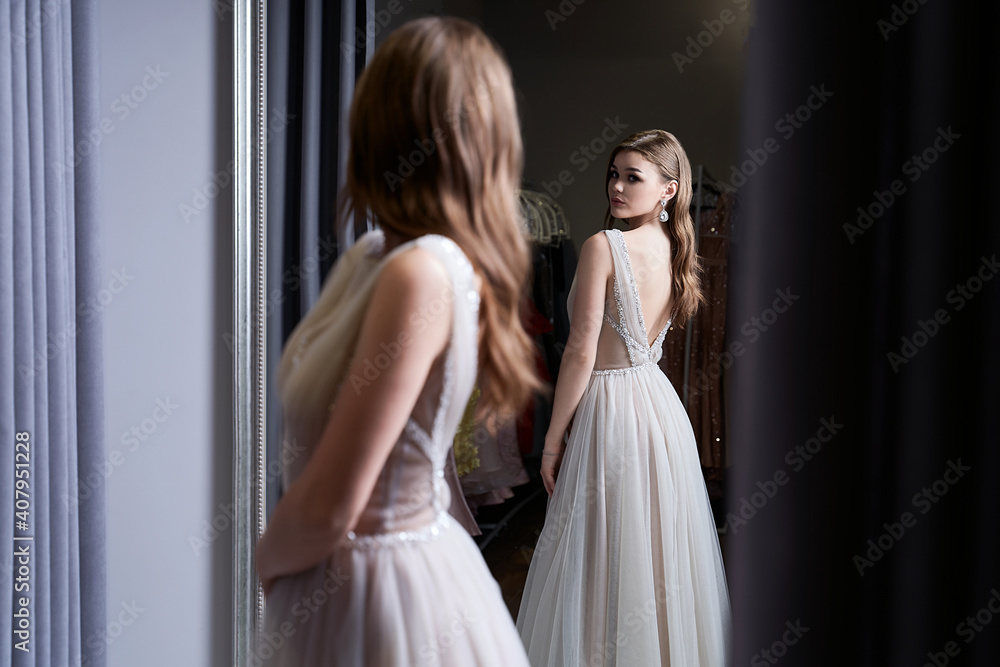 Young beautiful brunette girl wearing a full-length silver white chiffon prom ball gown decorated with sparkles and sequins. Model in front of mirror in a fitting room at dress hire service.