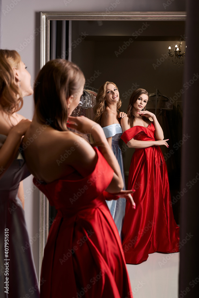 Two young beautiful girls wearing off-the-shoulder full-length sky blue and crimson red satin slit prom ball gowns. Models looking in mirror. Fitting room in dress hire service. Two young beautiful girls wearing off-the-shoulder full-length sky blue and crimson red satin slit prom ball gowns. Models looking in mirror. Fitting room in dress hire service.