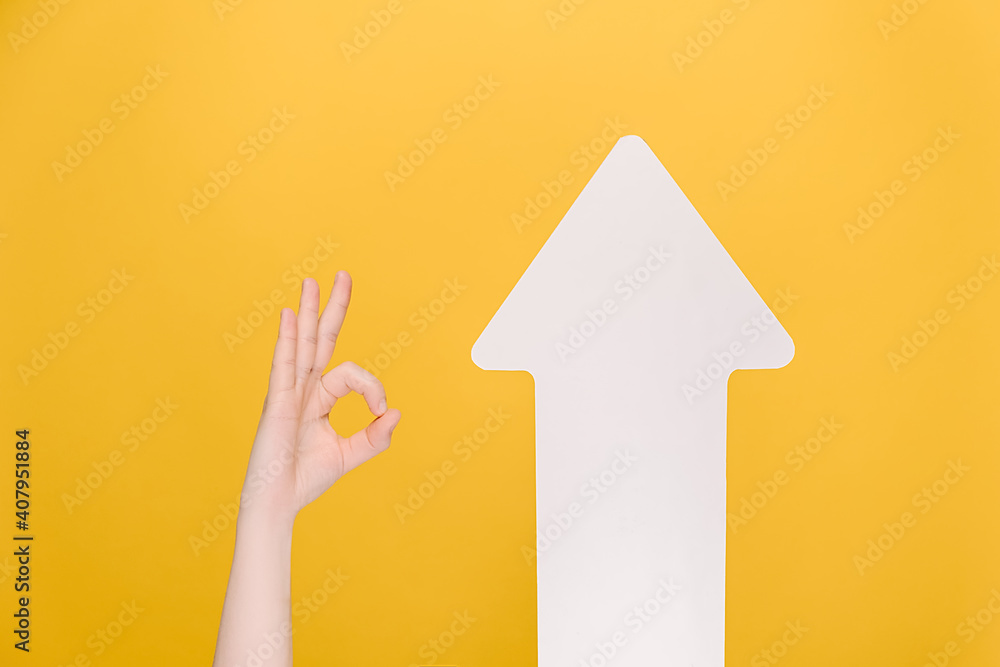 Naklejka premium Image of unrecognizable female makes thumb up gesture near white paper arrow, demonstrates approval or agreement, isolated on yellow background. Body language concept. Hand sign. Like gesture