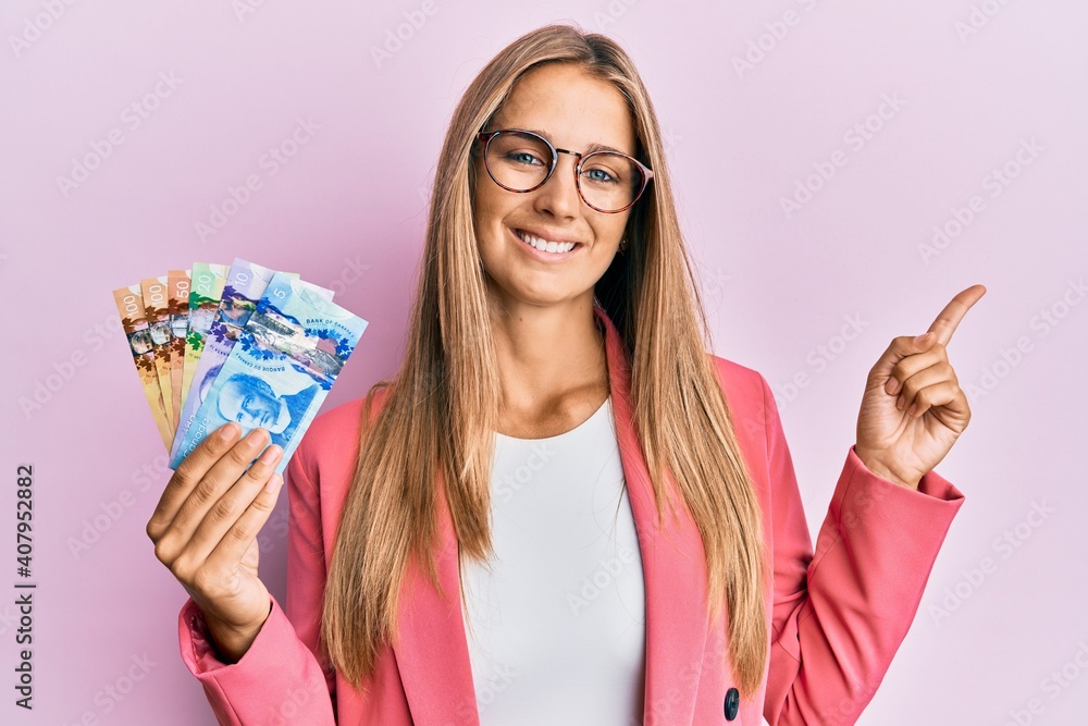 Obraz premium Young blonde woman wearing business style holding canadian dollars smiling happy pointing with hand and finger to the side
