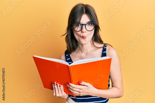 Young beautiful caucasian girl reading a book wearing glasses making fish face with mouth and squinting eyes, crazy and comical.
