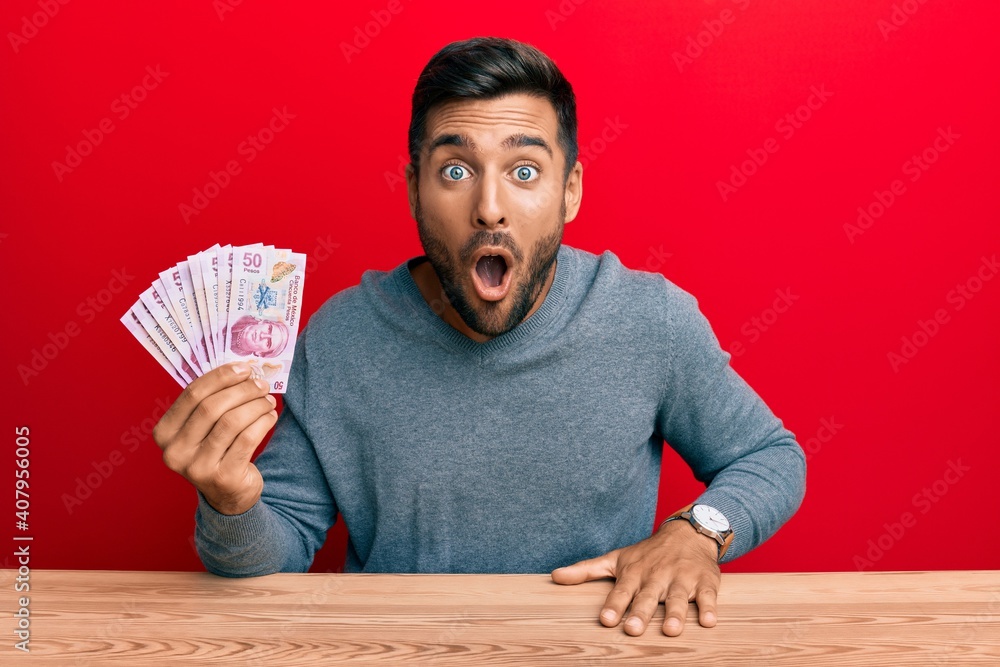 Handsome hispanic man holding mexican pesos scared and amazed with open mouth for surprise, disbelief face Handsome hispanic man holding mexican pesos scared and amazed with open mouth for surprise, disbelief face