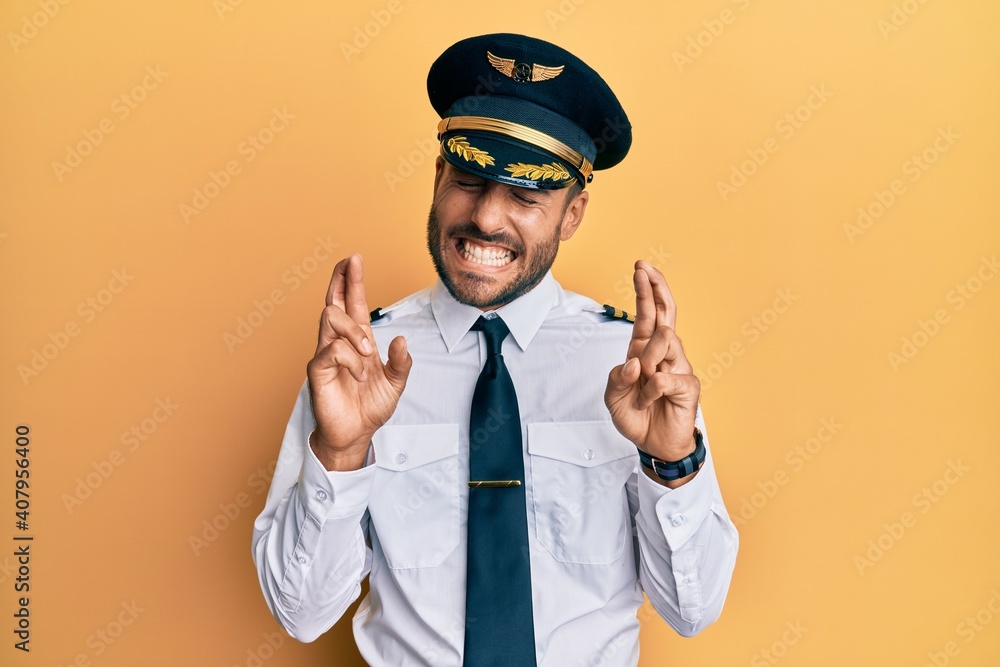 Handsome hispanic man wearing airplane pilot uniform gesturing finger crossed smiling with hope and eyes closed. luck and superstitious concept. Handsome hispanic man wearing airplane pilot uniform gesturing finger crossed smiling with hope and eyes closed. luck and superstitious concept.