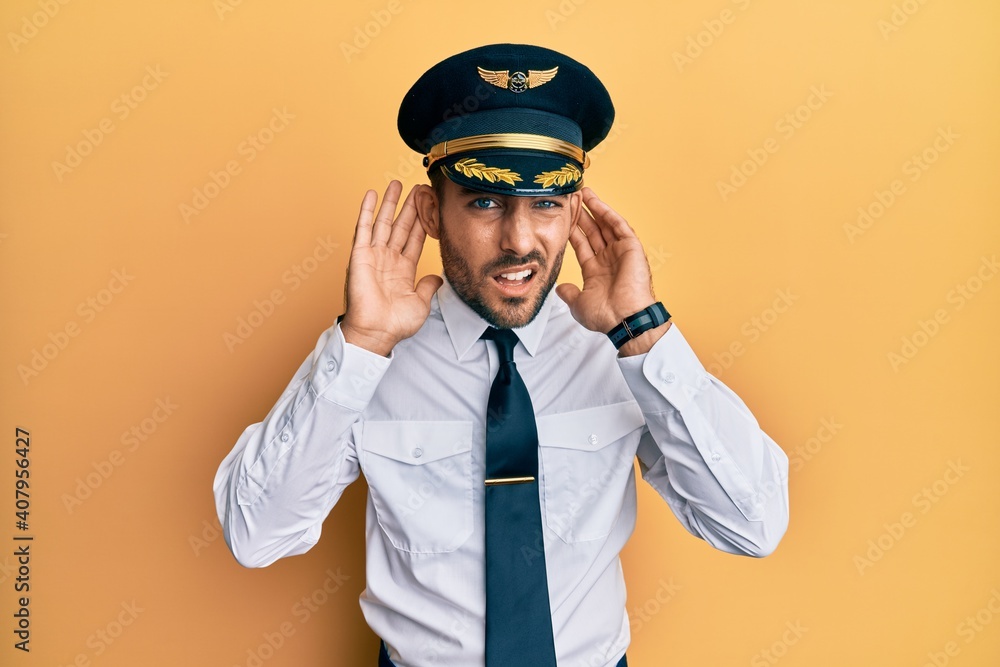 Handsome hispanic man wearing airplane pilot uniform trying to hear both hands on ear gesture, curious for gossip. hearing problem, deaf Handsome hispanic man wearing airplane pilot uniform trying to hear both hands on ear gesture, curious for gossip. hearing problem, deaf