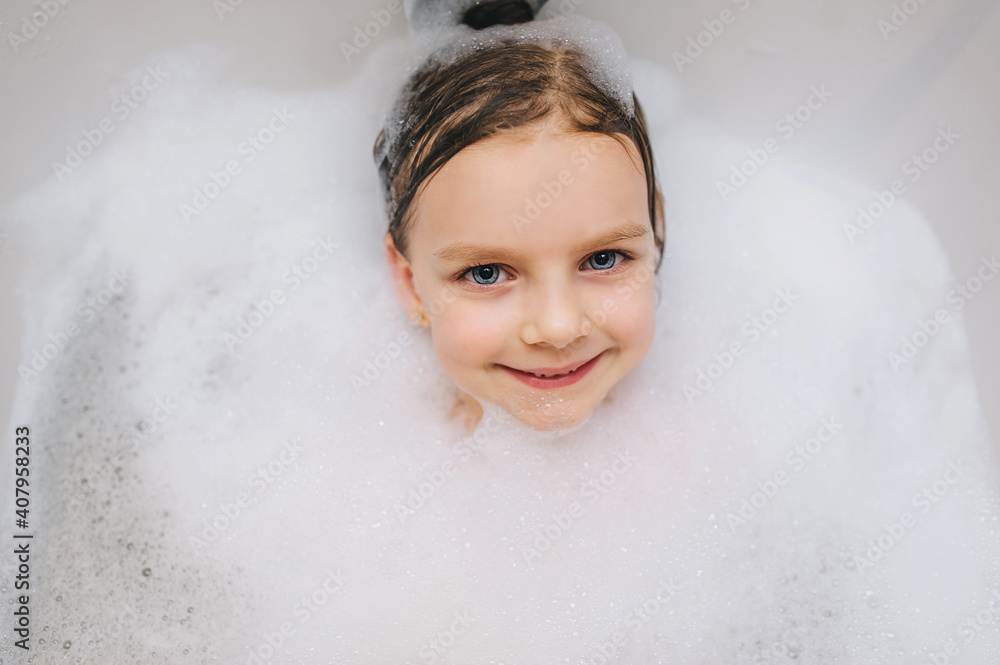 Close-up of a child's face. A little girl with long hair washes herself lying in a white bath with foam on her head from soap and shampoo. Model photography. Close-up of a child's face. A little girl with long hair washes herself lying in a white bath with foam on her head from soap and shampoo. Model photography.