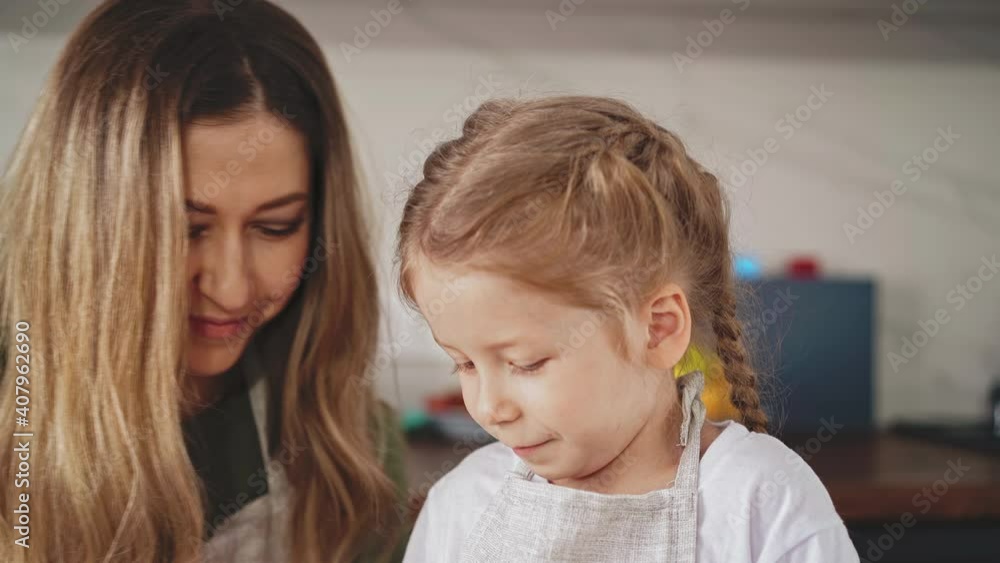 crane shot, close up: young mother and little girl with long hair are in kitchen in gray aprons, cook food together, smile at each other, against background of different fruits and vegetables crane shot, close up: young mother and little girl with long hair are in kitchen in gray aprons, cook food together, smile at each other, against background of different fruits and vegetables