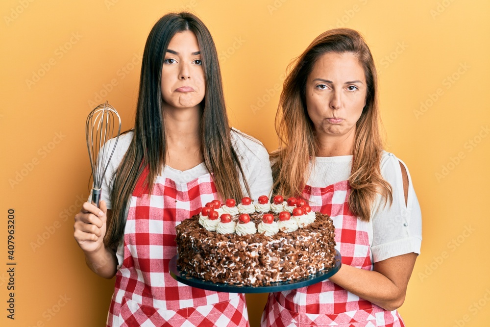 Hispanic family of mother and daughter wearing baker apron holding homemade cake depressed and worry for distress, crying angry and afraid. sad expression.