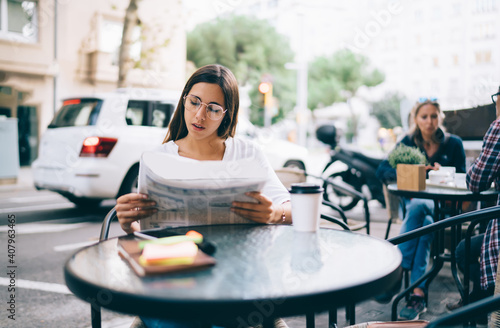 Serious caucasian woman in eyewear for vision correction sitting at table on cfe terrace reading morning news in daily printed media, pensive female reading holding newspaper searching vacancy photo