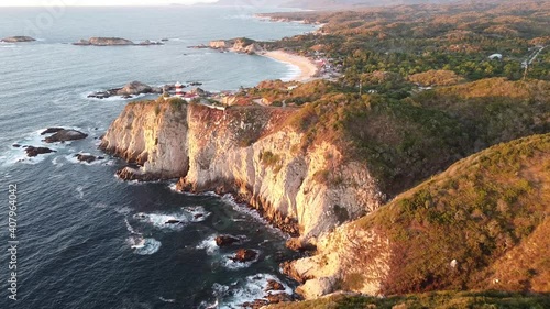 Vista aérea en reversa de un acantilado donde se ve un faro  y una playa a lo lejos.  photo