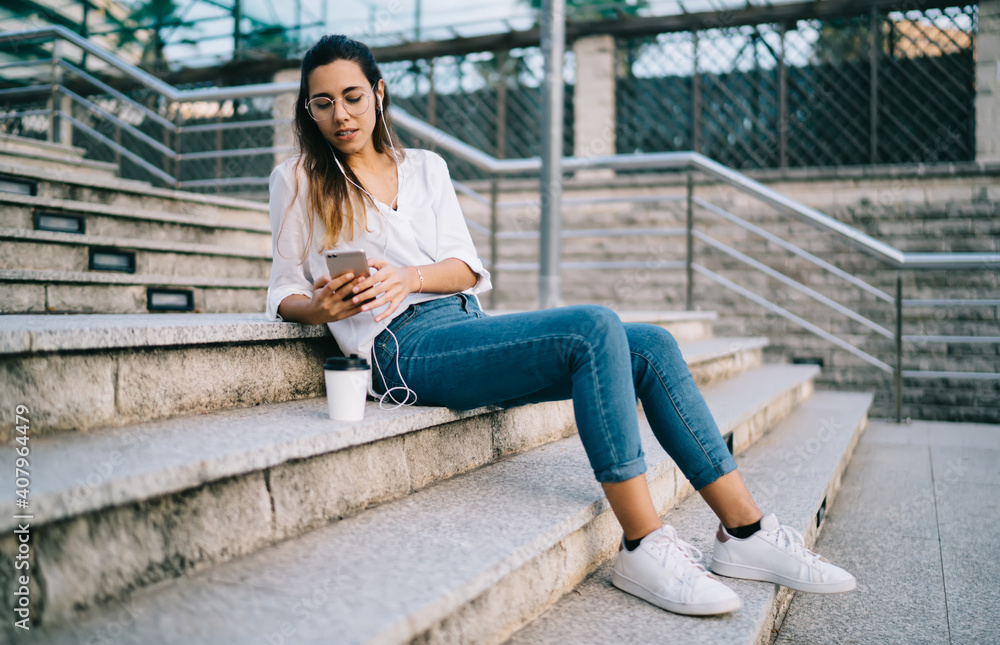 Serious caucasian woman in earphones choosing radio for listening music on mobile phone recreating on urban settings, pensive hipster girl watching vlog on smartphone having coffee break outdoors Serious caucasian woman in earphones choosing radio for listening music on mobile phone recreating on urban settings, pensive hipster girl watching vlog on smartphone having coffee break outdoors