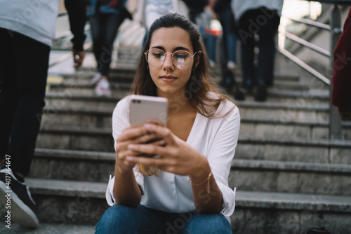 Beautiful caucasian woman in eyewear sitting outdoors on crowded city stairs holding mobile phone watching video online from vlog,charming hipster girl millennial sending messages and text on cellular