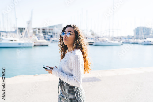 Youthful Turkish tourist in optical eyewear feeling pondering during travel promenade for exploring city holding modern cellphone device in hands, Hispanic hipster girl with smartphone tech © GalakticDreamer