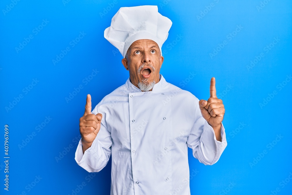 Middle age grey-haired man wearing professional cook uniform and hat amazed and surprised looking up and pointing with fingers and raised arms. Middle age grey-haired man wearing professional cook uniform and hat amazed and surprised looking up and pointing with fingers and raised arms.