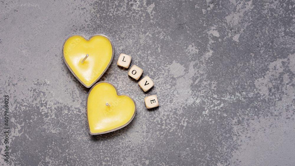 Obraz premium Greeting card for Valentine day. Two Yellow candles in heart shape on trendy grey backround. Text in English LOVE on wooden cubes. color 2021