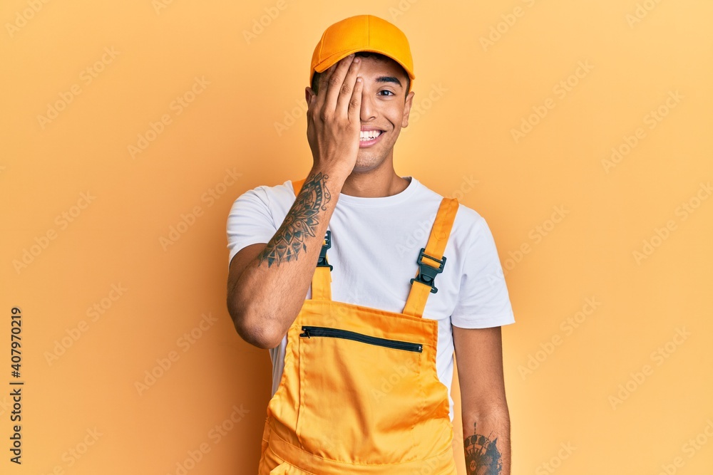 Young handsome african american man wearing handyman uniform over yellow background covering one eye with hand, confident smile on face and surprise emotion. Young handsome african american man wearing handyman uniform over yellow background covering one eye with hand, confident smile on face and surprise emotion.