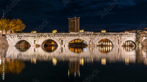 Ponte romano antico sopra al fiume di notte con grattacielo sullo sfondo e luci situato a Rimini