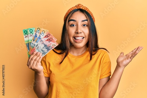 Young latin woman holding australian dollars banknotes celebrating achievement with happy smile and winner expression with raised hand