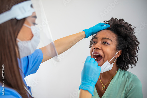 Professional medical worker wearing personal protective equipment testing woman for dangerous disease using test stick. Woman opens mouth for cheek and throat swab while being tested for Covid-19