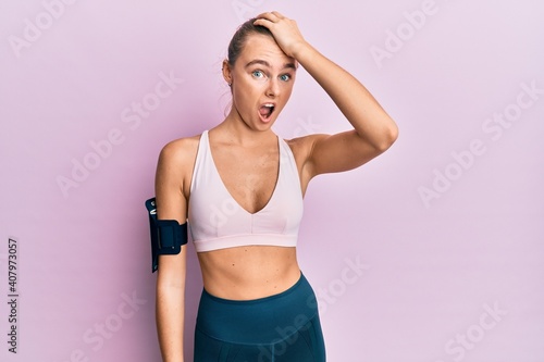 Beautiful blonde woman wearing sportswear and arm band surprised with hand on head for mistake  remember error. forgot  bad memory concept.