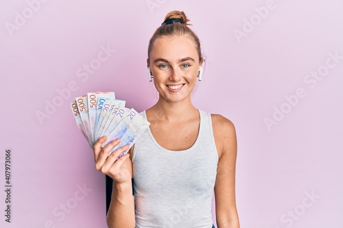 Beautiful blonde sport woman holding 20 swedish krona banknotes looking positive and happy standing and smiling with a confident smile showing teeth