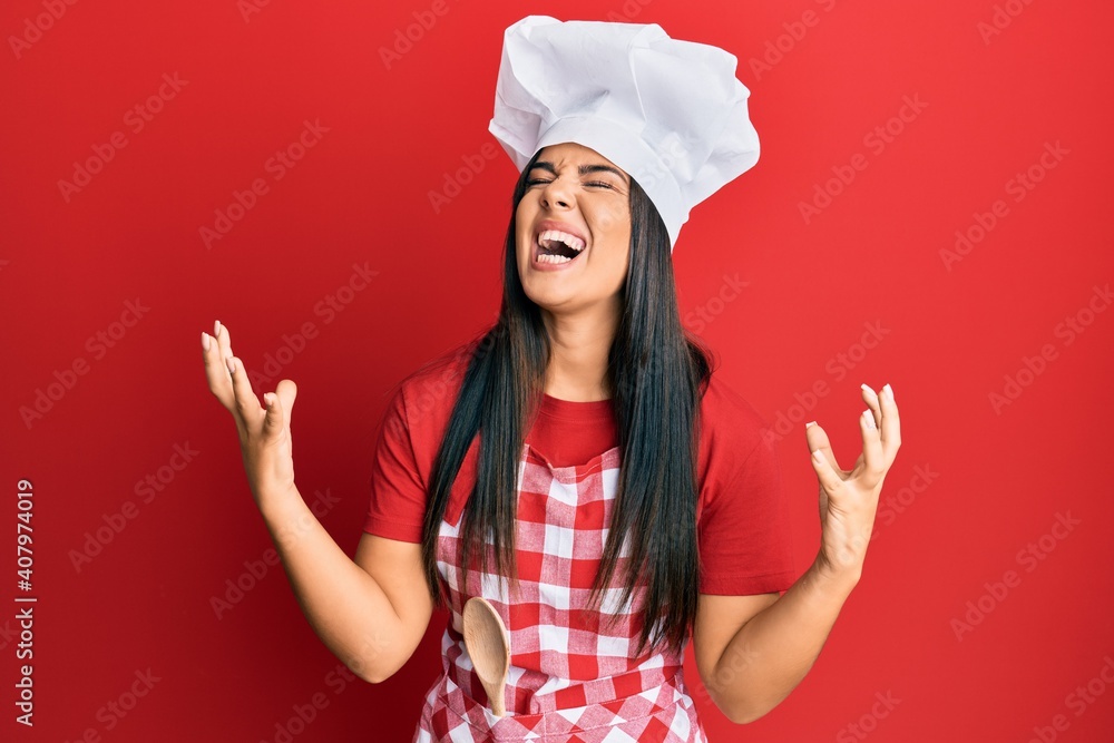 Young beautiful hispanic girl wearing baker uniform and cook hat crazy and mad shouting and yelling with aggressive expression and arms raised. frustration concept. Young beautiful hispanic girl wearing baker uniform and cook hat crazy and mad shouting and yelling with aggressive expression and arms raised. frustration concept.