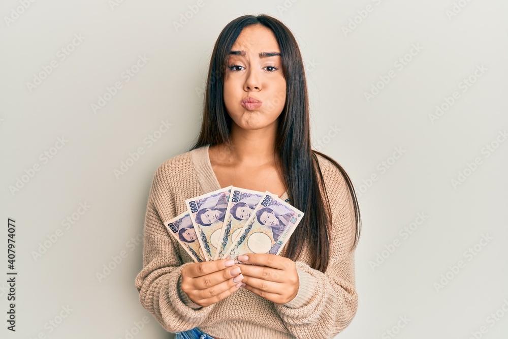 Young beautiful hispanic girl holding japanese yen banknotes puffing cheeks with funny face. mouth inflated with air, catching air. Young beautiful hispanic girl holding japanese yen banknotes puffing cheeks with funny face. mouth inflated with air, catching air.