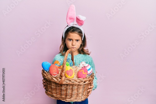 Little beautiful girl wearing cute easter bunny ears holding wicker basket with colored eggs skeptic and nervous, frowning upset because of problem. negative person.