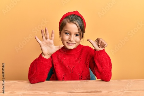 Little beautiful girl wearing casual clothes sitting on the table showing and pointing up with fingers number six while smiling confident and happy.