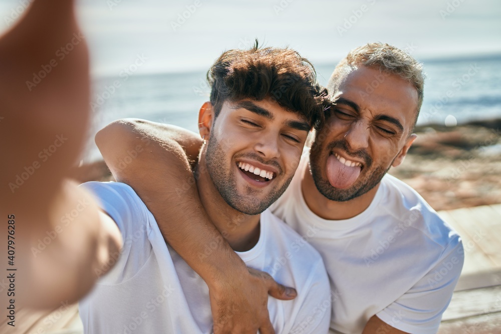 Fototapeta premium Young gay couple smiling happy making selfie by the smartphone at the beach.