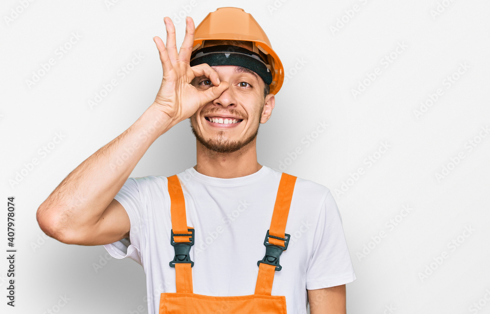 Hispanic young man wearing handyman uniform and safety hardhat smiling happy doing ok sign with hand on eye looking through fingers