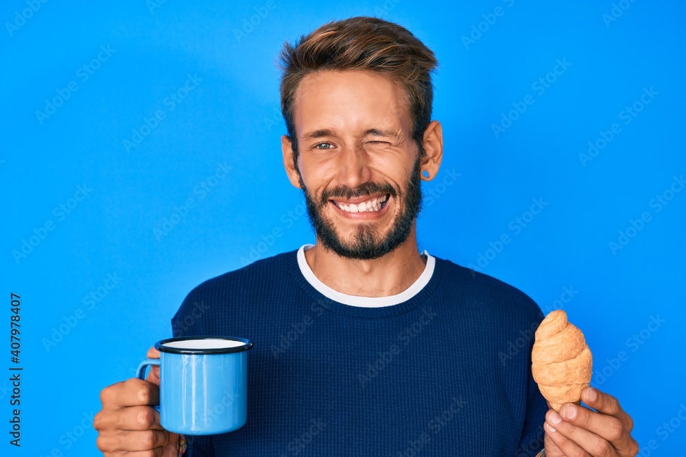 Handsome caucasian man with beard drinking a cup of coffee and croissant winking looking at the camera with sexy expression, cheerful and happy face. Handsome caucasian man with beard drinking a cup of coffee and croissant winking looking at the camera with sexy expression, cheerful and happy face.