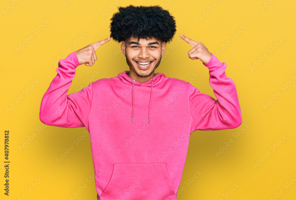 Young african american man with afro hair wearing casual pink sweatshirt smiling pointing to head with both hands finger, great idea or thought, good memory Young african american man with afro hair wearing casual pink sweatshirt smiling pointing to head with both hands finger, great idea or thought, good memory