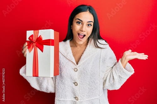 Young brunette woman holding gift celebrating achievement with happy smile and winner expression with raised hand