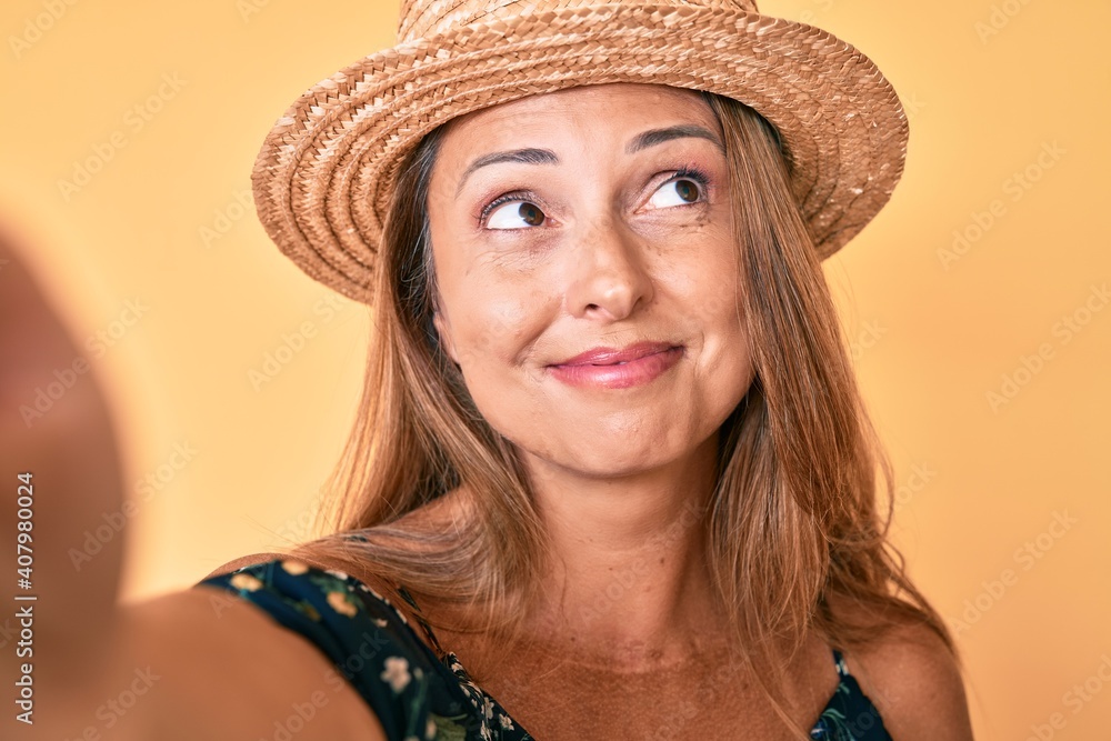 Middle age hispanic woman wearing summer hat taking a selfie smiling looking to the side and staring away thinking. Middle age hispanic woman wearing summer hat taking a selfie smiling looking to the side and staring away thinking.
