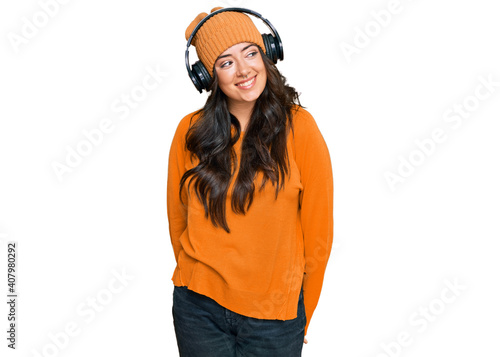 Beautiful brunette young woman listening to music using headphones looking away to side with smile on face, natural expression. laughing confident.
