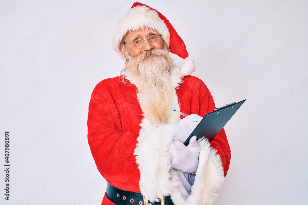 Old senior man with grey hair and long beard wearing santa claus costume writting list smiling with a happy and cool smile on face. showing teeth. Old senior man with grey hair and long beard wearing santa claus costume writting list smiling with a happy and cool smile on face. showing teeth.