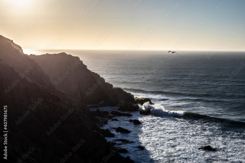 An awe sunset in an idyllic beach landscape with the sunset over the water and the sunlight generating a moody atmosphere. An amazing wil scenery with the cliffs and the sea waves splashing water