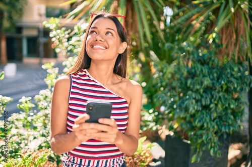 Young latin girl smiling happy using smartphone at the city.