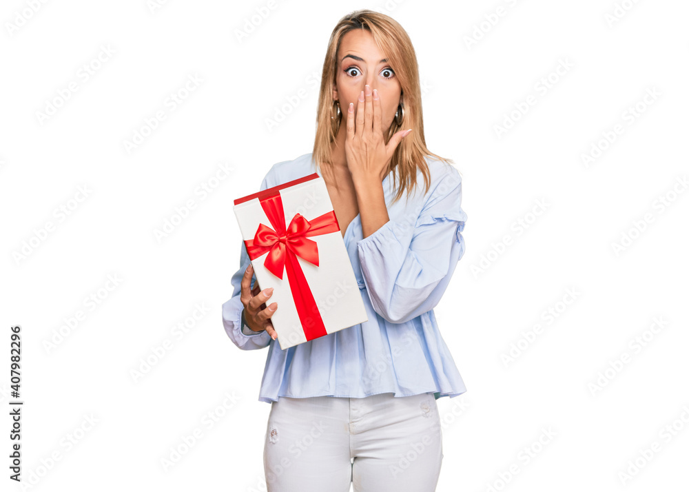 Beautiful blonde woman holding gift covering mouth with hand, shocked and afraid for mistake. surprised expression Beautiful blonde woman holding gift covering mouth with hand, shocked and afraid for mistake. surprised expression