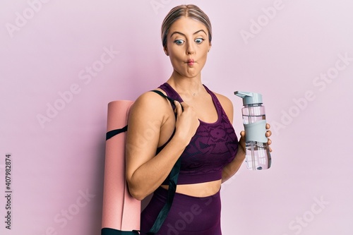 Beautiful blonde woman wearing sportswear holding water bottle and yoga mat making fish face with mouth and squinting eyes, crazy and comical.